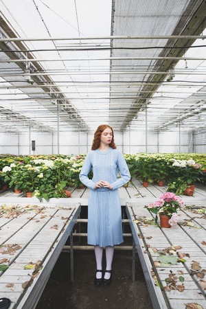 Vertical shot of redhead pretty girl between half-empty tables looking at flowers in orangeryの写真素材