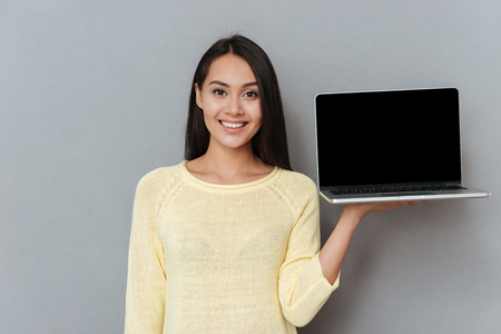 Happy lovely young woman holding blank screen laptop over grey backgroundの写真素材