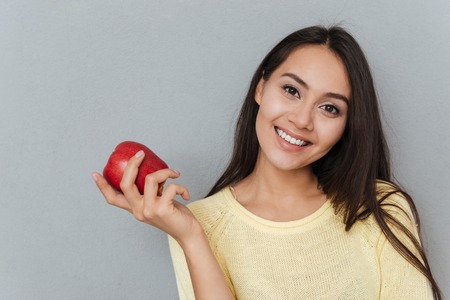 Cheerful charming young woman holding red apple over grey backgroundの写真素材