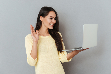 Cheerful charming young woman holding laptop and having video conference over grey backgroundの写真素材