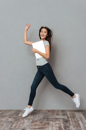 Happy pretty young woman holding laptop and running over grey backgroundの写真素材