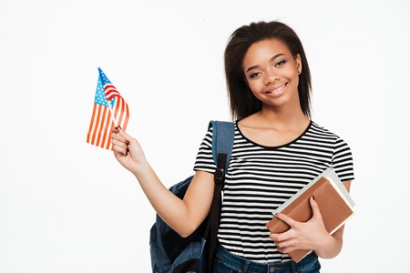 Portrait of a happy afro american girl student with backpack holding books and US flag isolated over white backgroundの写真素材