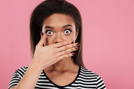 Close up portrait of a shocked african girl looking at camera and covering her mouth with hand isolated over pink backgroundの写真素材