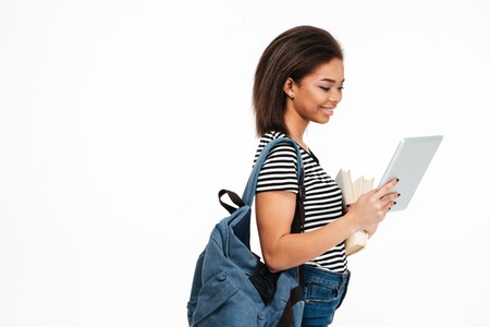 Portrait of a smiling cute african student girl with backpack using pc tablet isolated over white...