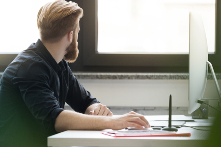 Side view of a bearded young man sitting at his desk and gazing out a windowの写真素材