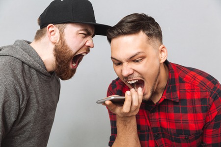 Picture of screaming young two men standing over grey wall and talking by phone. Looking aside.