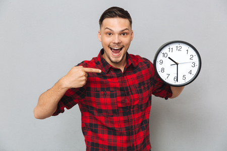 Happy man in shirt holding clock and pointing at their over gray backgroundの写真素材