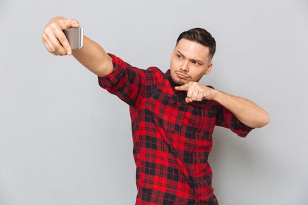 Handsome confident man in plaid shirt taking selfie on smartphone isolated on gray background