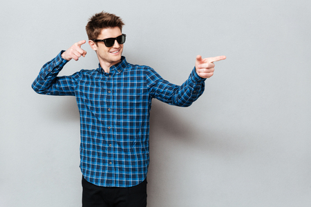 Image of young happy man wearing sunglasses standing over grey wall and looking aside while pointing.の写真素材