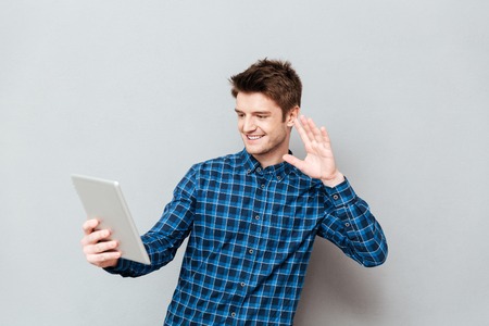 Image of happy young man standing over grey wall and talking with friends by tablet computer while waving. Looking aside.の写真素材