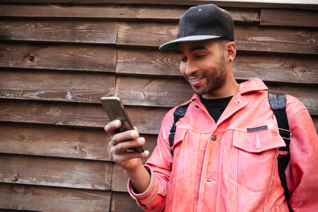 Portrait of a happy smiling afro american man in hat typing message on mobile phone while standing...