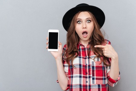 Portrait of a shocked casual girl in plaid shirt pointing finger at blank screen mobile phone isolated over gray backgroundの写真素材