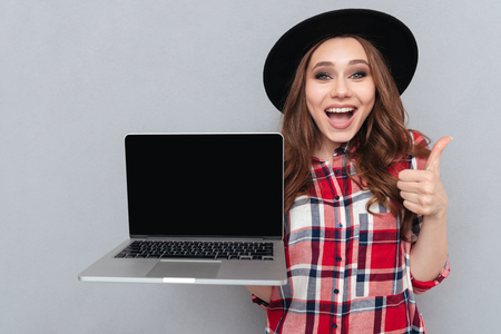 Portrait of a smiling happy girl in plaid shirt holding blank screen laptop computer and showing thumbs up gesture isolated over gray backgroundの写真素材