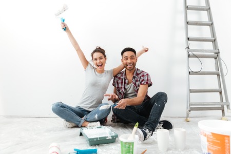 Photo of young happy loving couple sitting on floor in new flat makes repair. Looking at camera.の写真素材