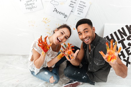 Cheerful young couple with hands dirty in colorful paints sitting over white backgroundの写真素材