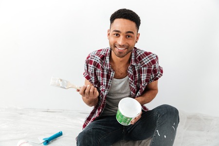 Portrait of afro american cheerful man sitting on floor with repair equipment and dye isolatedの写真素材