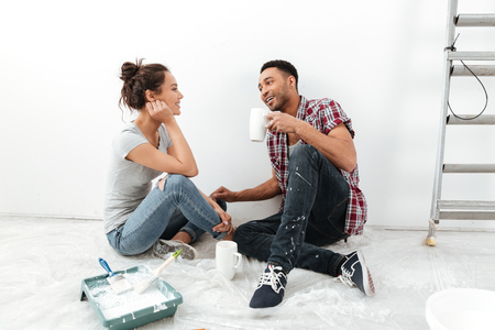 Picture of smiling young loving couple sitting on floor in new flat makes repair. Looking aside while drinking tea.の写真素材