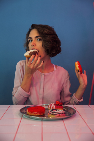 Hungry young girl eating donuts at the table indoors and looking at cameraの写真素材