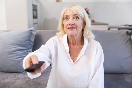 Happy senior woman watching tv and changing channels with remote control while sitting on a couch at...