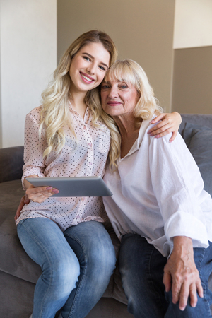 Smiling grandmother and her granddaughter holding a tablet and looking at vamera while sitting on a couch at homeの写真素材