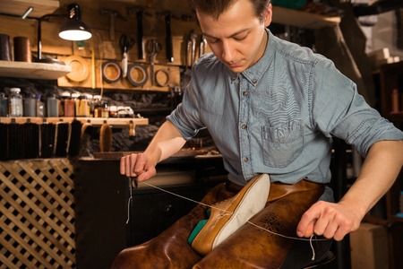 Picture of young concentrated shoemaker in workshop making shoes. Looking aside.