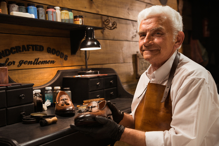 Image of mature smiling shoemaker in workshop making shoes. Looking at camera.