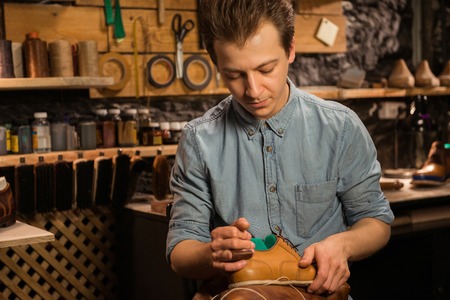 Photo of young handsome shoemaker sitting in workshop making shoes. Looking aside.