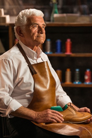 Image of mature handsome shoemaker in workshop holding shoes. Looking aside.の写真素材
