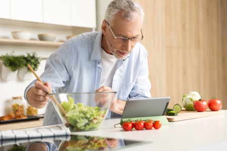 Photo of concentrated mature man wearing glasses standing at the kitchen cooking salad using tablet computer. Looking aside.の写真素材