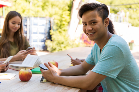 Photo of smiling young multiethnic friends students outdoors using mobile phones. Asian man looking camera.の写真素材