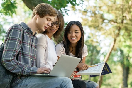 Photo of multiethnic group of young concentrated students sitting and studying outdoors while using laptop. Looking aside.の写真素材
