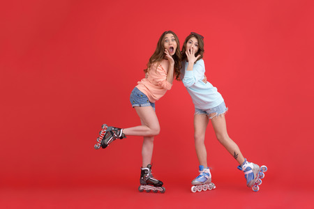 Picture of young shocked ladies standing on rollers isolated over red background. Looking at camera.