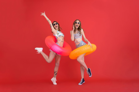 Picture of amazing young ladies dressed in swimwear with rubber rings jumping isolated over red background. Looking camera.の写真素材