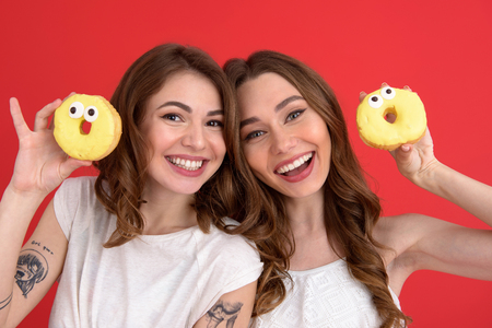 Image of young smiling two ladies friends standing with donuts isolated over red background. Looking at camera.の写真素材