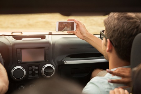 Young man taking a selfie with smartphone while sitting at the front seat in a car with his friends...
