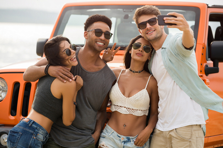 Group of young cheerful friends talking a selfie together while standing outdoors with a car on a backgroundの写真素材