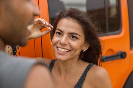 Close up of a smiling young woman talking to a guy while standing near a car outdoors
