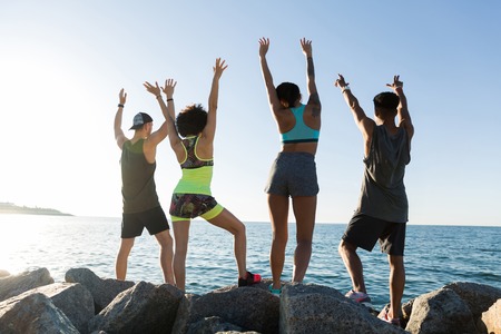 Back view of a group of happy sports people standing at the beach side with hands raisedの写真素材