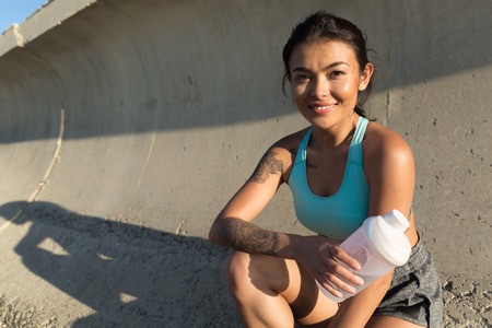 Beautiful smiling sporty woman sitting and resting with bottle of water outdoorsの写真素材