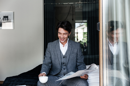 Happy smiling businessman reading newspaper and drinking coffee at hotel roomの写真素材