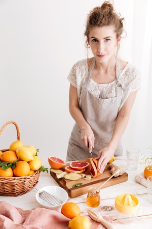 Young cheerful lady standing indoors near table with a lot of citruses and cut the grapefruit....