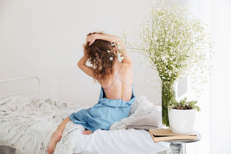 Back view picture of amazing young woman sitting indoors on bed stretching. Focus on flowers.の写真素材
