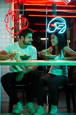 Image of young multiethnic cheerful loving couple sitting in cafe looking aside eating.の写真素材