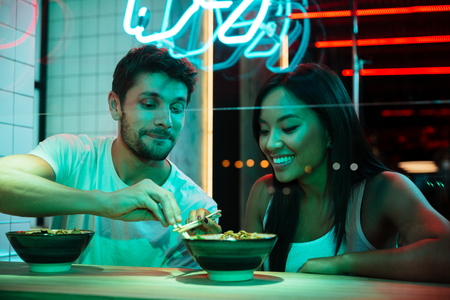 Picture of young multiethnic smiling loving couple sitting in cafe looking aside eating.