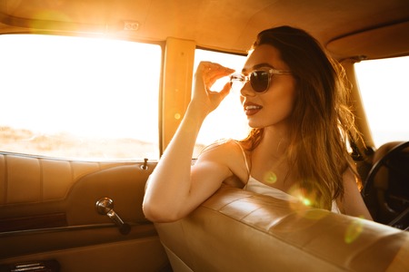 Beautiful young woman in sunglasses sitting on a front seat inside a car and looking backwards