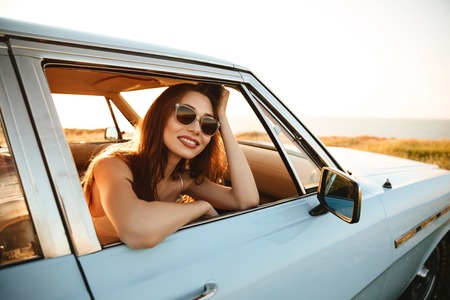 Happy young woman in sunglasses leaning on a window and looking away while sitting inside a carの写真素材