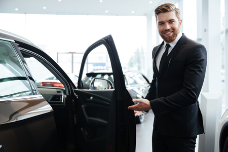 Happy smiling male dealer inviting customers to sit in a new carの写真素材