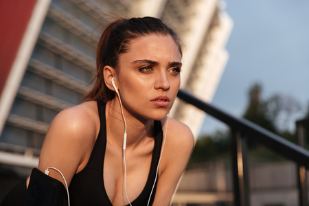 Picture of concentrated serious young sports woman sitting with earphones outdoors and looking aside.の写真素材