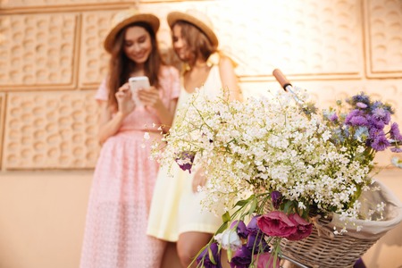 Image of two young happy ladies standing outdoors using phone near bicycle. Looking aside. Focus on flowers.の写真素材