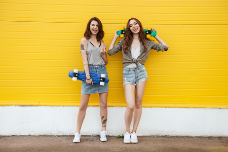 Image of two young happy women friends standing over yellow wall. Looking at camera holding skateboards.の写真素材